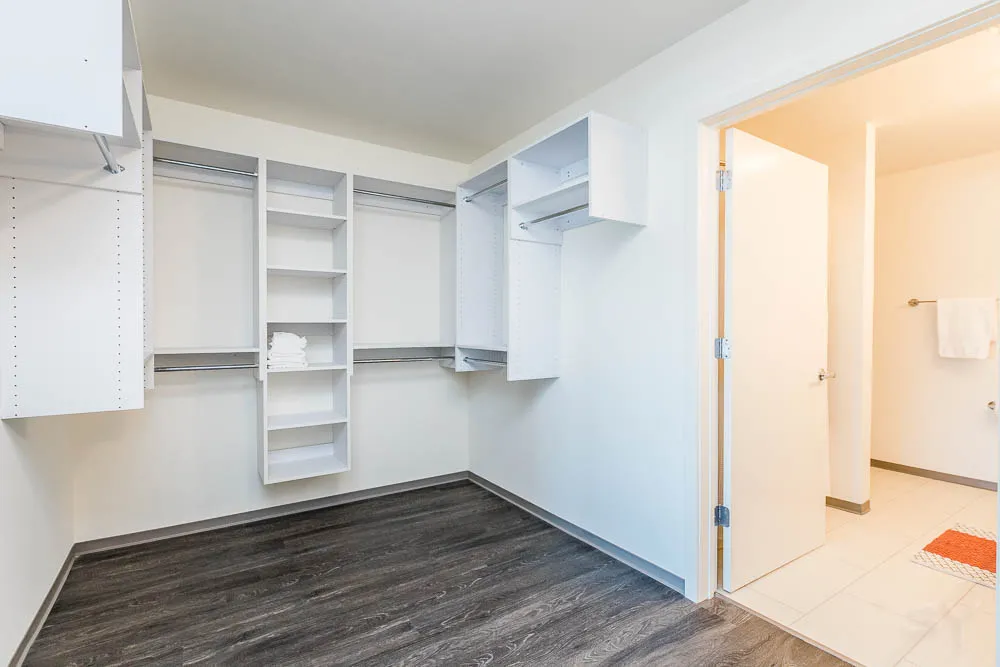 Spacious empty walk-in closet with white shelves and hanging rods, wooden floor, and an open door leading to a lit bathroom.
