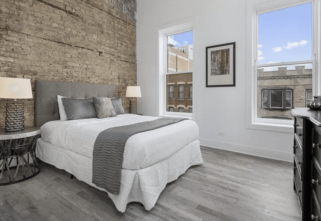 A modern bedroom with a brick accent wall, a bed with gray and white bedding, two side tables with lamps, and two large windows overlooking a building.