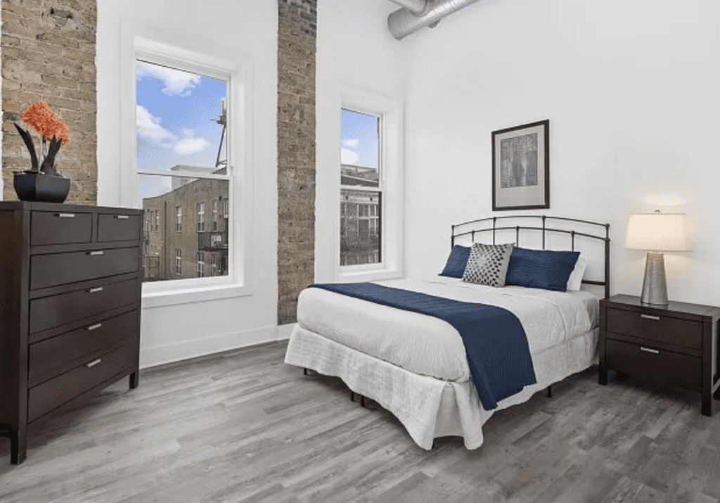 Bright bedroom with a metal-framed bed, blue bedding, wooden dresser, and nightstand with a lamp. Exposed brick wall and large windows provide city views. Light gray wood floor.