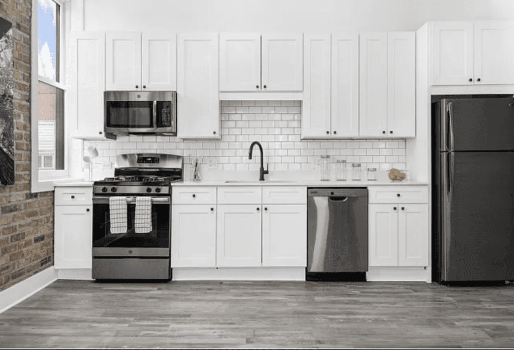Modern kitchen with white cabinets, stainless steel appliances including a fridge, oven, microwave, and dishwasher, brick accent wall, and a white subway tile backsplash.