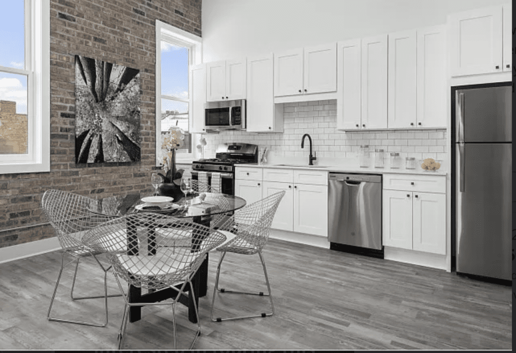 Modern kitchen with white cabinets, stainless steel appliances, brick accent wall, and round glass dining table with wire chairs. A black and white abstract painting hangs on the wall.