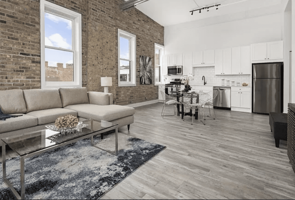 Spacious open-plan living area with brick walls, gray sofa, glass coffee table, dining set, and modern kitchen featuring white cabinets and stainless steel appliances.
