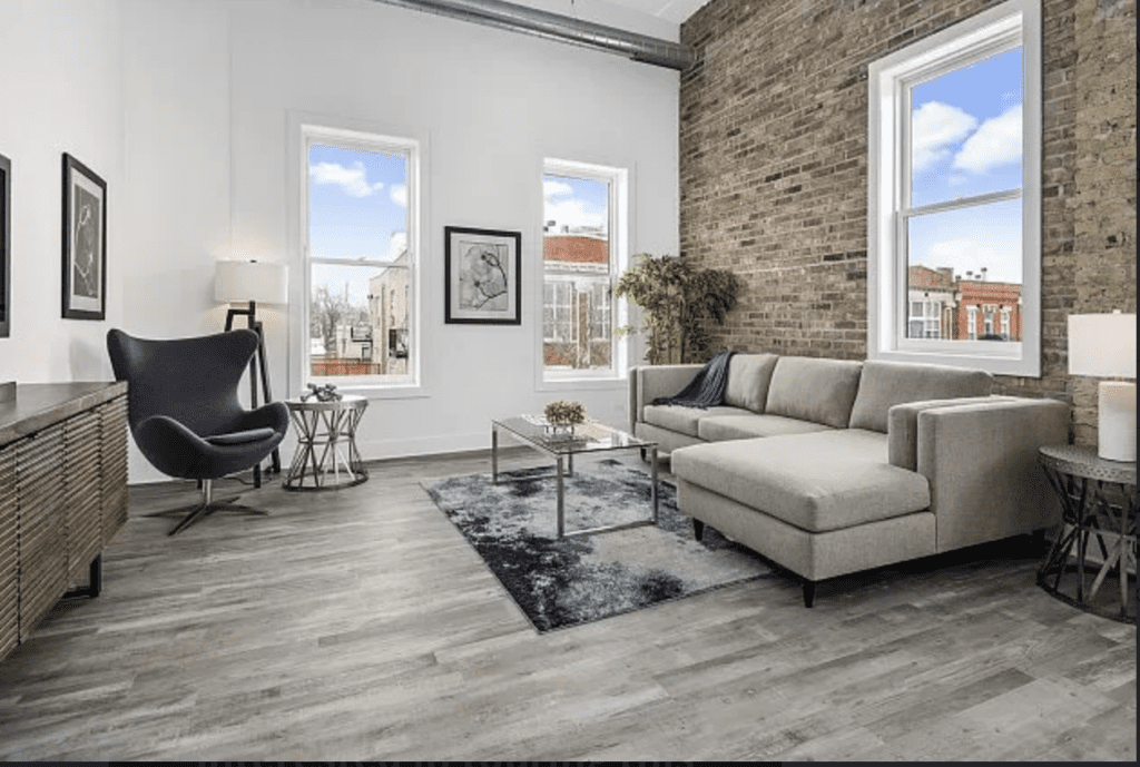 Modern living room with a gray sectional sofa, brick accent wall, armchair, coffee table, and abstract art. Large windows with city views; light wood flooring.