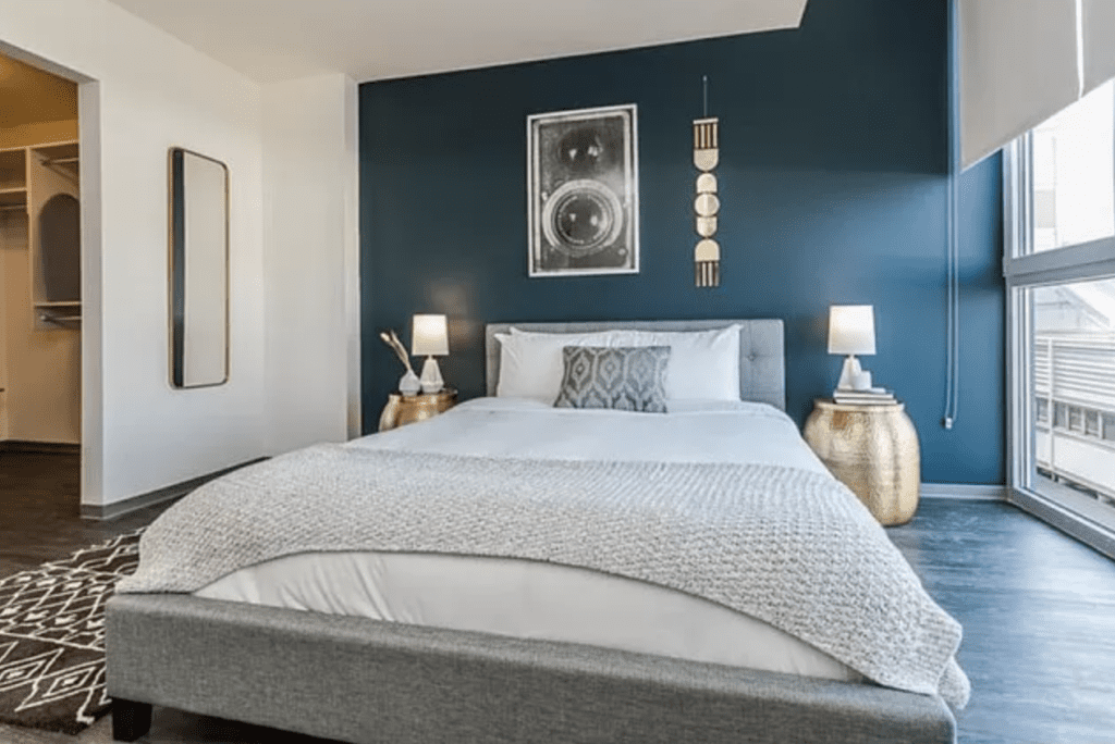 Bedroom with a gray upholstered bed, blue accent wall, decorative art, two gold side tables with lamps, a patterned rug, and a closet on the left. Large window on the right side.