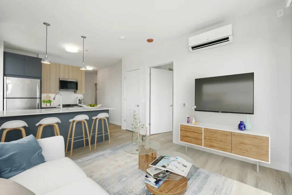 Modern living room with an open kitchen. Features a sofa, wall-mounted TV, round coffee table with magazines, kitchen island with stools, and a stainless steel fridge. Light wood and white decor.