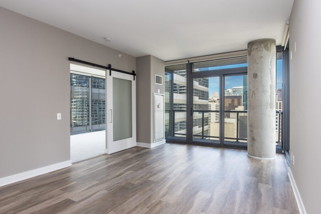 Empty modern apartment with large windows, sliding door, and a view of the city. Light wood flooring and a concrete pillar are visible.