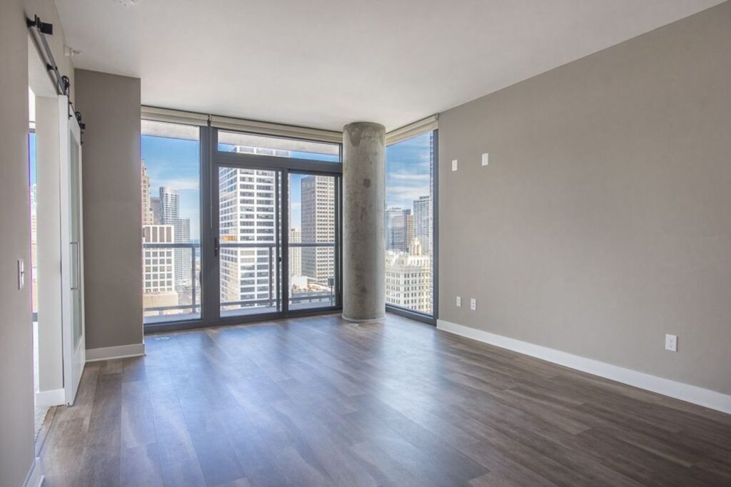 Empty modern apartment with large windows, wood flooring, and a view of city skyscrapers. Gray walls and a visible concrete pillar add to the contemporary design.