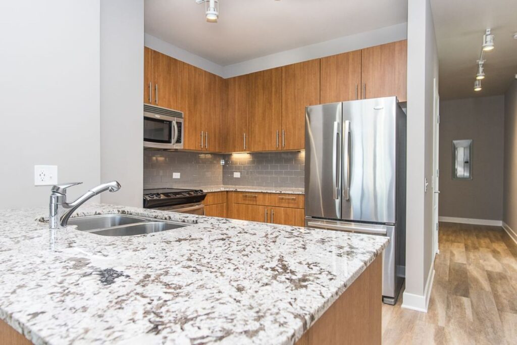 Modern kitchen with granite countertops, stainless steel appliances, including a fridge and oven, wooden cabinets, and light wood flooring.