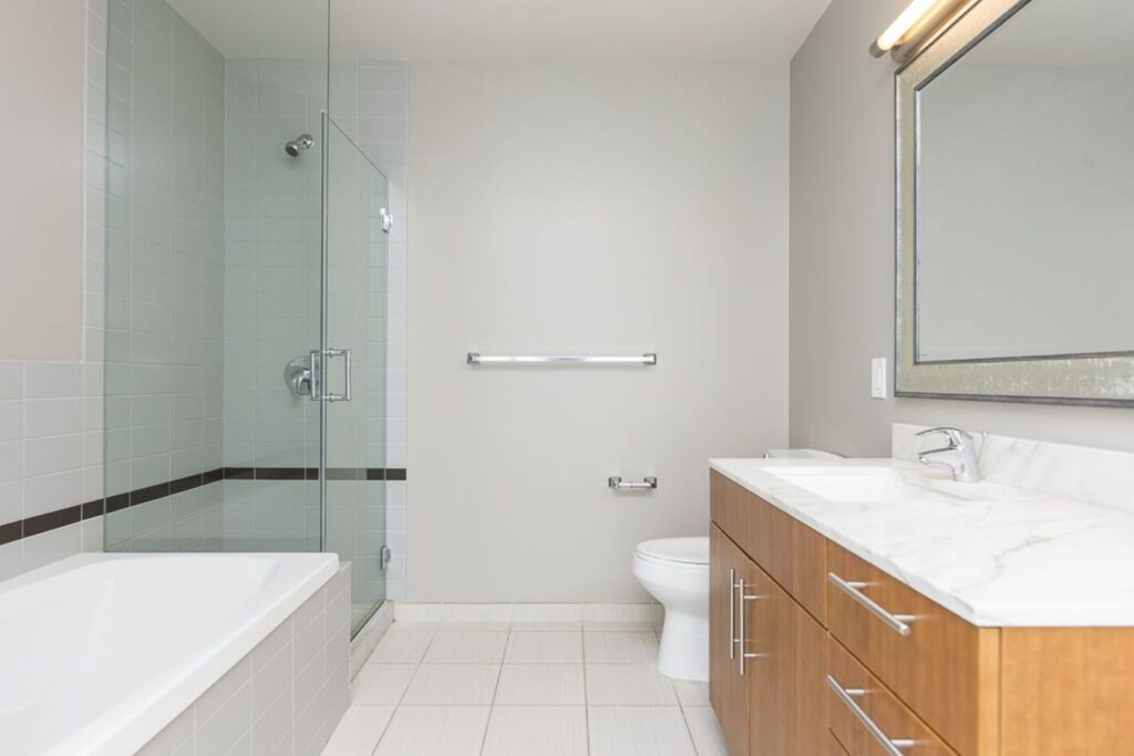 Modern bathroom with glass shower, bathtub, toilet, and wooden vanity with marble countertop. Large mirror above sink, neutral-toned tiles, and recessed lighting.
