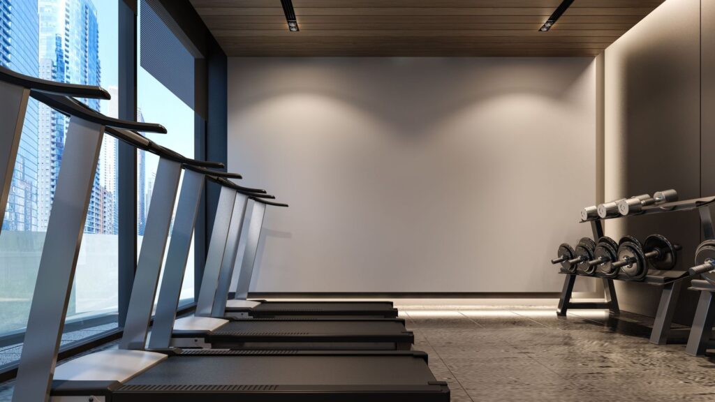 Empty gym room with several treadmills on the left, weight bench and dumbbells on the right, and large windows showing city buildings.