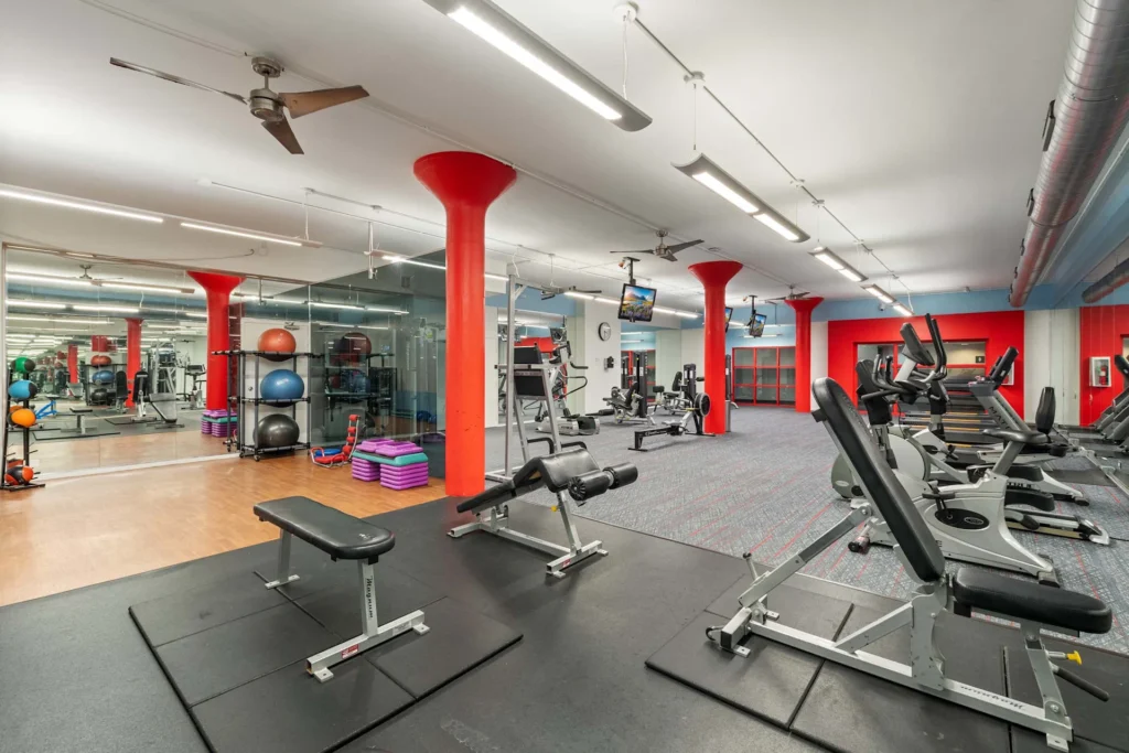 A spacious gym features treadmills, weight benches, exercise balls, and fitness equipment against red pillars and mirrored walls.