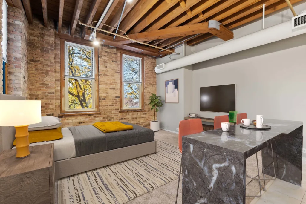 Loft-style bedroom with exposed brick walls and wooden beams, featuring a bed, TV, and a marble dining table with red chairs. A small plant and a wall art piece decorate the space.