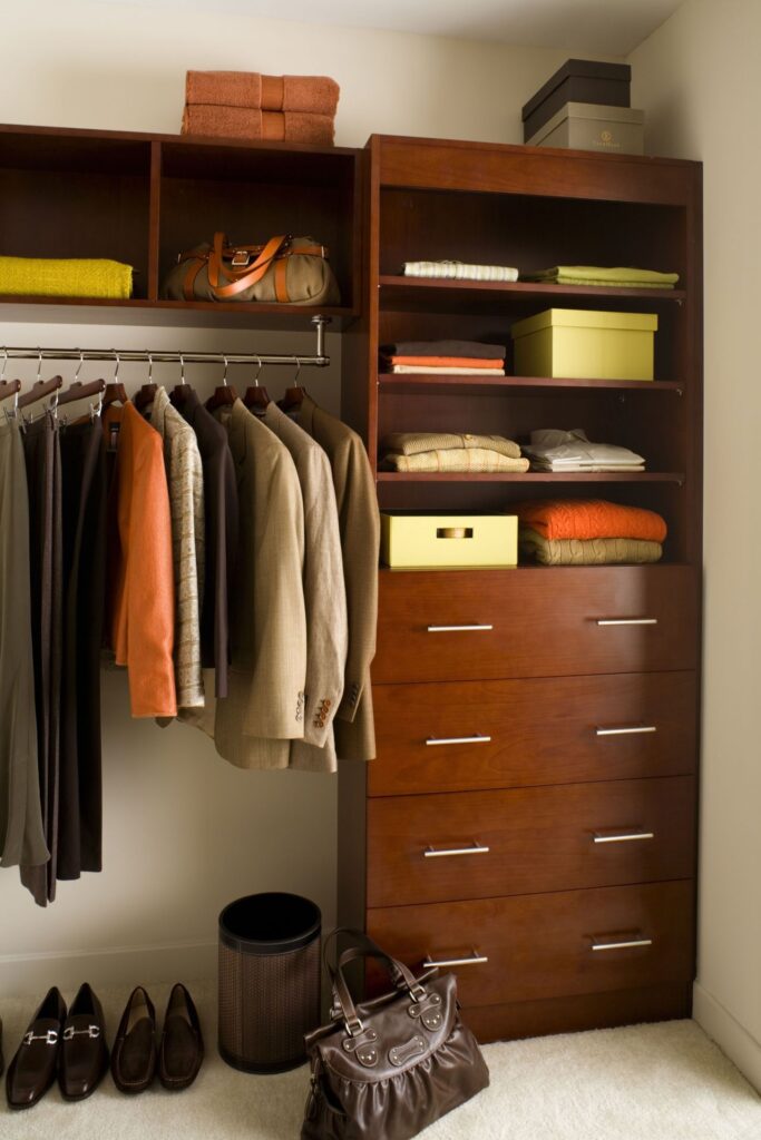 A wooden closet with hanging jackets, organized shelves with boxes, folded clothes, and a row of shoes. A handbag is placed on the floor beside a trash bin.