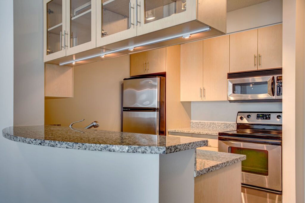 Modern kitchen with stainless steel appliances, light wood cabinets, and granite countertops; features an electric stove, refrigerator, and a curved countertop with overhead cabinets.