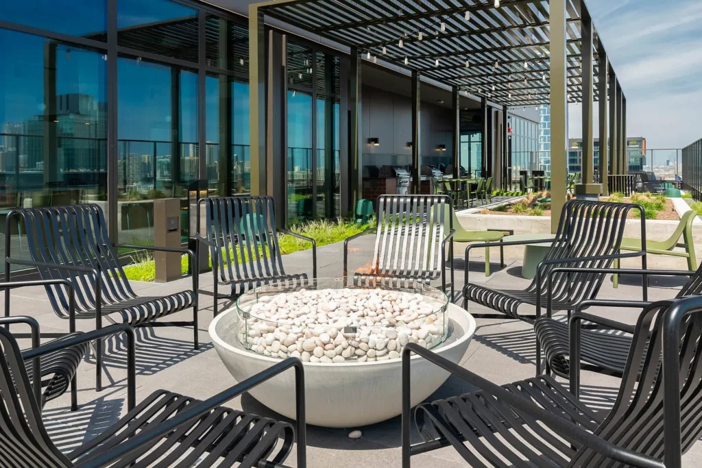 Rooftop patio with a circular fire pit surrounded by black metal chairs. Glass windows reflect the city skyline. Overhead pergola provides partial shade.