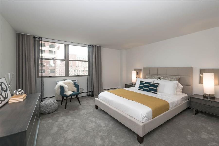 Modern bedroom with a large bed featuring a mustard-colored throw, two nightstands with lamps, a window with city views, and an accent chair with a faux fur throw.