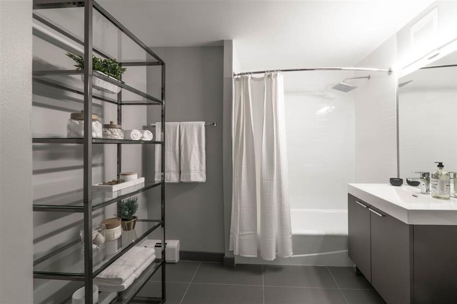 Modern bathroom with gray tile flooring, a white shower curtain, and a dark metal shelf stocked with towels and toiletries. A long mirror hangs above a sink with a white countertop.