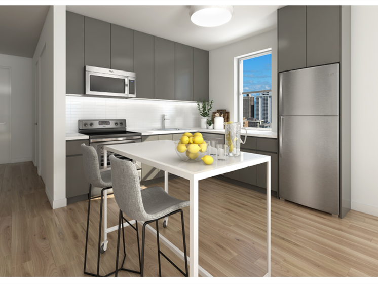 Modern kitchen with gray cabinets, stainless steel appliances, and a minimalist table with gray chairs. A bowl of lemons and a pitcher are on the table. Window view shows a cityscape.