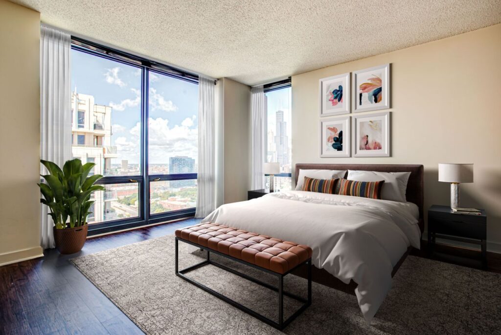 Bedroom interior with large window view, featuring a bed with white bedding, a bench at the foot, framed artwork above, a potted plant to the left, and a lamp on a nightstand to the right.