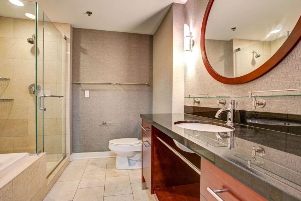 Modern bathroom with a glass-enclosed shower, a round mirror above a dark granite countertop with a sink, and a toilet beside neutral-colored walls and tiled flooring.