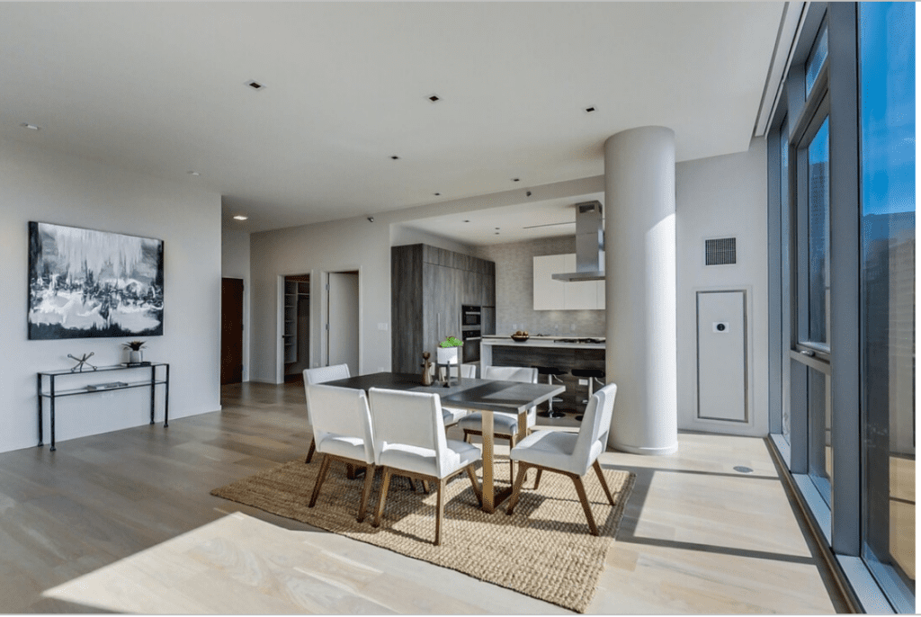 Modern open-plan living area with large windows, light wood floors, a dining table with six chairs on a rug, and a kitchen with gray cabinetry and stainless steel appliances.