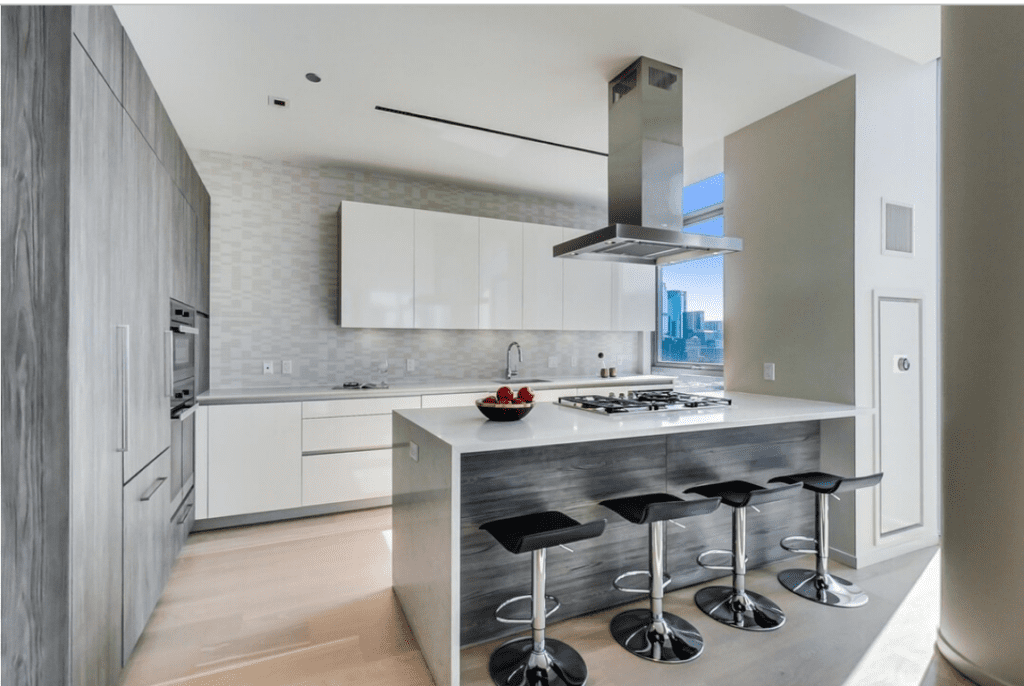Modern kitchen with gray and white cabinets, island with stools, stainless steel appliances, and a bowl of apples. Large windows with a cityscape view.
