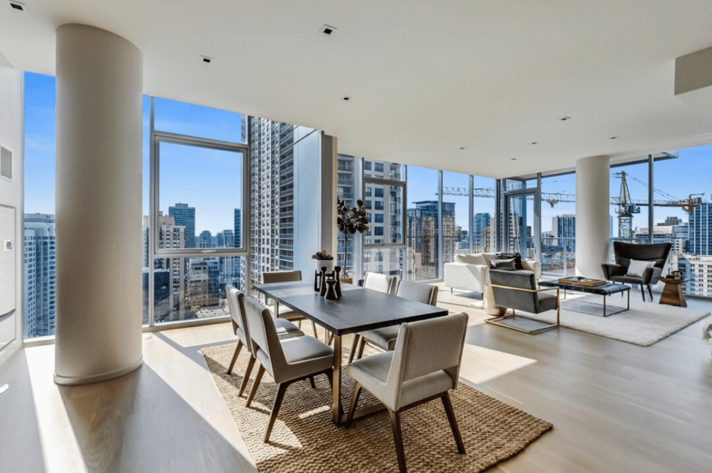 Modern open-plan apartment with floor-to-ceiling windows, city view, dining table, chairs, sofas, and a coffee table on a light wood floor.