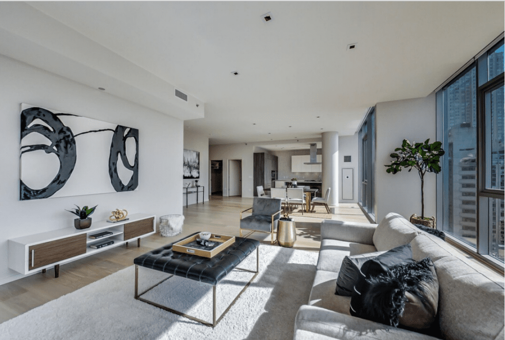 Spacious modern living room with large windows, light wood floors, a sectional sofa, abstract wall art, and a view into a kitchen with a dining area.