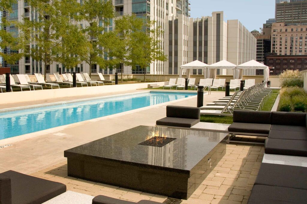 Outdoor pool area with lounge chairs, cabanas, and a fire pit, surrounded by city buildings.