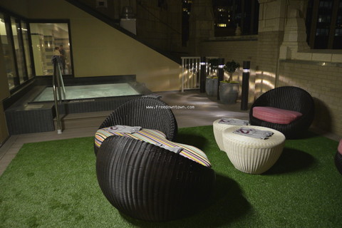 Outdoor seating area with round black chairs and white tables on artificial grass, near a hot tub. White wall and glass doors in the background.