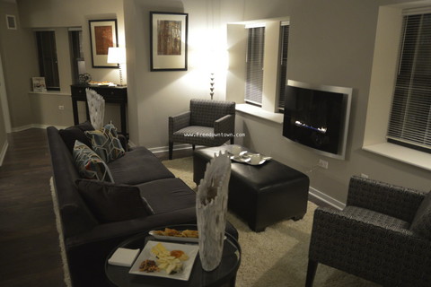 Modern living room with dark sofas, armchairs, a wall-mounted TV, and a lit fireplace. A small table holds snacks and decor. Artwork hangs on light gray walls.