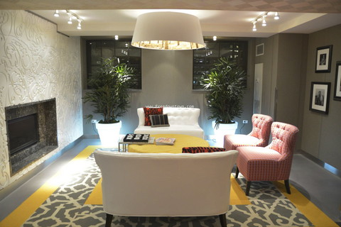 Modern living room with a white sofa, two pink chairs, a yellow ottoman, and a fireplace. Two large plants are near the windows, with a rug and overhead lighting completing the decor.