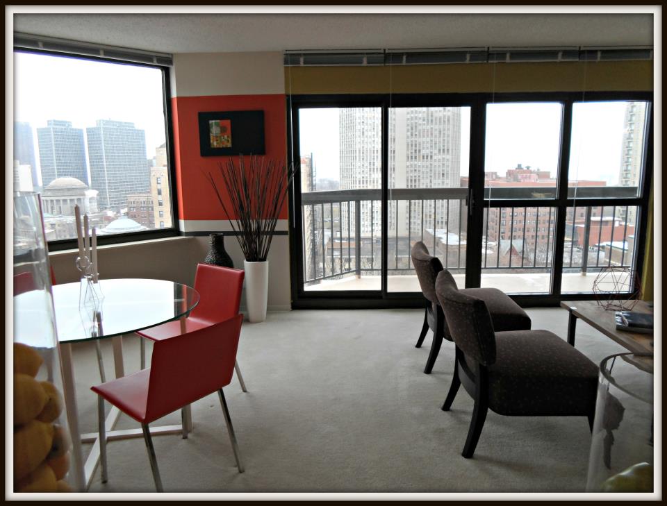 Modern apartment interior with large windows, dining table, red chair, and cityscape view. Neutral-colored chairs face the windows, and contemporary decor accents the space.