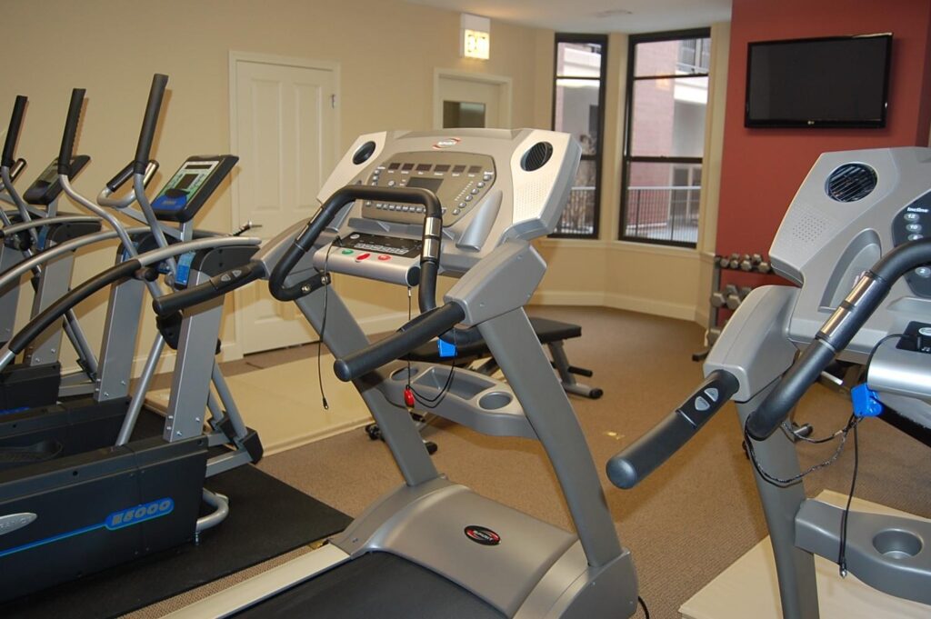 A gym with treadmills, ellipticals, and a wall-mounted TV. Barbell weights are visible in a corner on a rack. The room has a window showing an exterior building view.