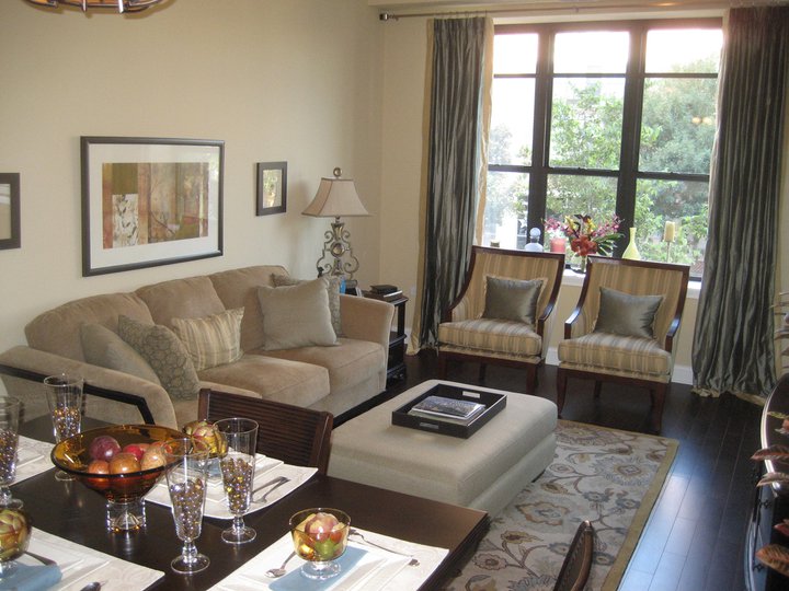 Stylish living room with a sofa, two chairs, and a large window. Dining table set with plates and glasses. Art on the walls, table with fruit bowl, and an upholstered ottoman in the center.