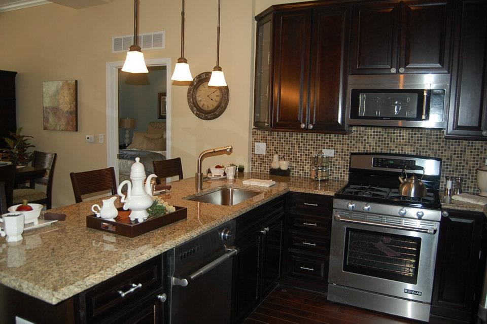 Modern kitchen with dark wood cabinets, granite countertops, and stainless steel appliances. Central island with teapot set. Wall clock and mosaic tile backsplash visible. Hanging lights above.