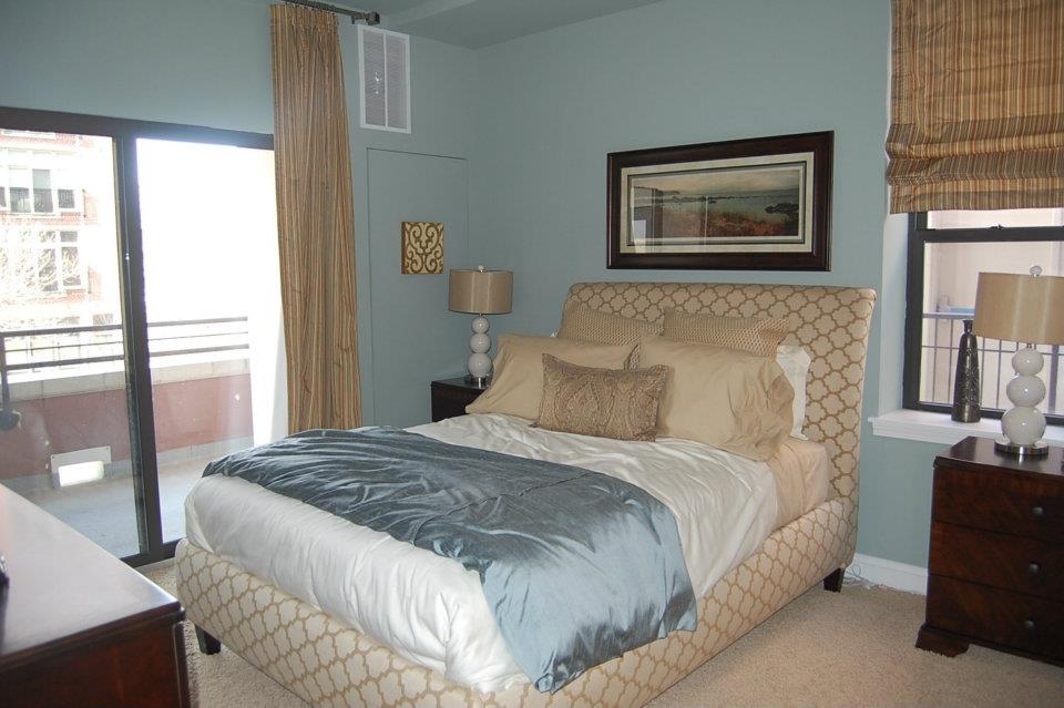 Bedroom with a patterned bed, blue throw, two side tables with lamps, framed art on the wall, and a window with beige curtains leading to a balcony.