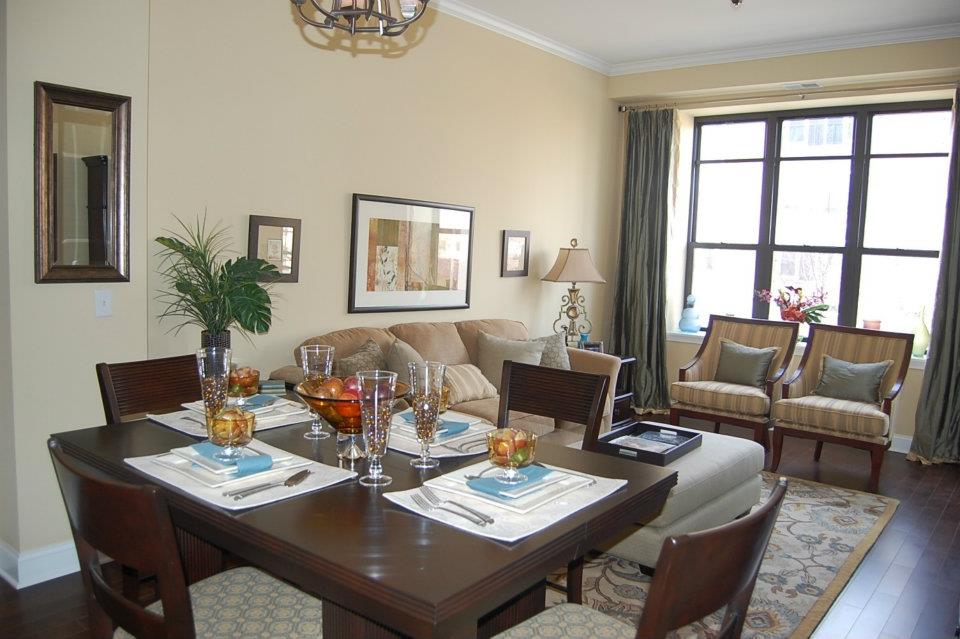 Dining area with a wooden table set for four, adjacent to a living room with a sofa and armchairs. Large windows with curtains allow natural light. Decor includes framed art and indoor plants.
