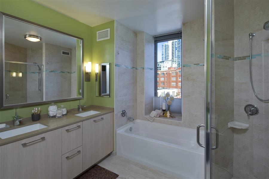 Modern bathroom with a double vanity, bathtub, and glass-enclosed shower. Light green and beige decor. Window with a city view.