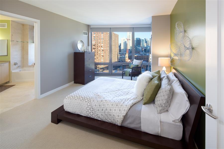 Modern bedroom with green accent wall, large bed, white and gray bedding, dark wood furniture, and city view through large windows. A bathroom is partially visible on the left.