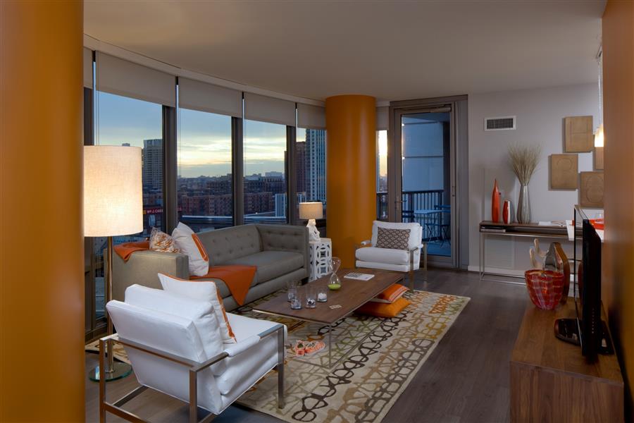 Modern living room with orange columns, large windows, and city view. Features gray sofa, white chairs, wooden coffee table, and patterned rug.