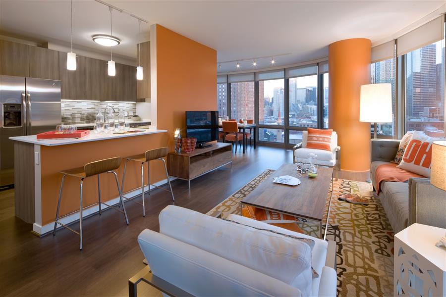 Modern living room with orange and beige decor, featuring a kitchen with bar stools, a sofa, armchairs, a TV, and large windows overlooking a cityscape.