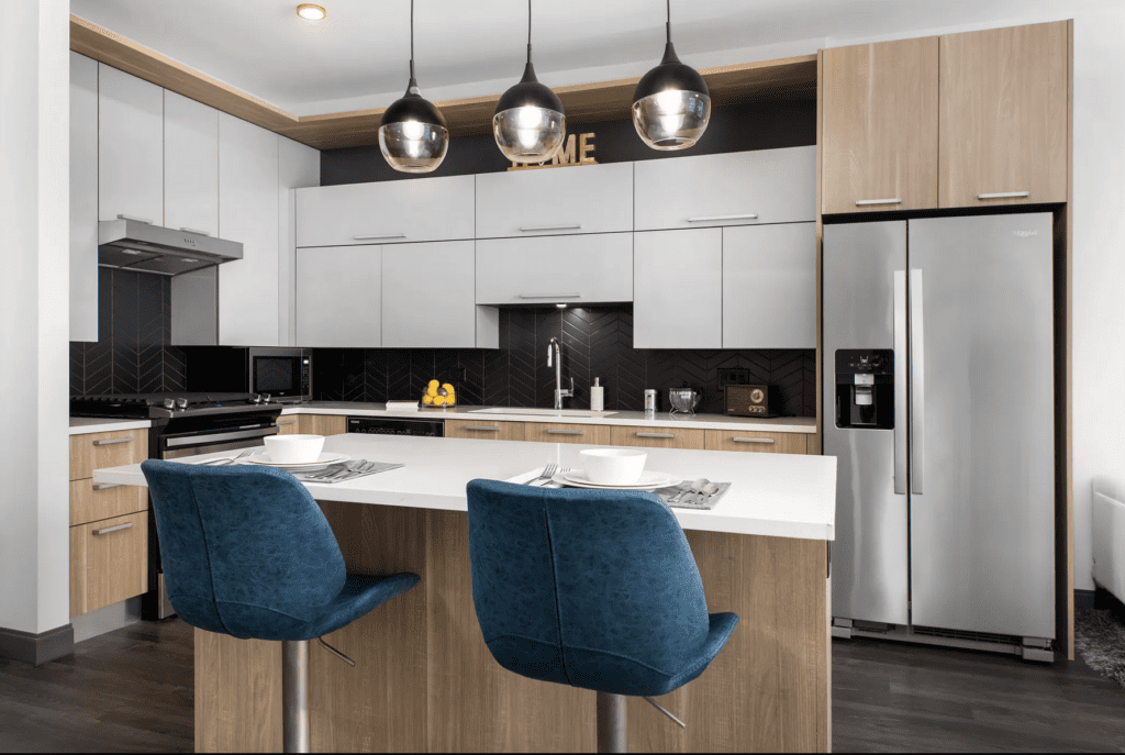 Modern kitchen with wood and white cabinets, a stainless steel fridge, black herringbone backsplash, and a center island with two blue chairs under pendant lights.