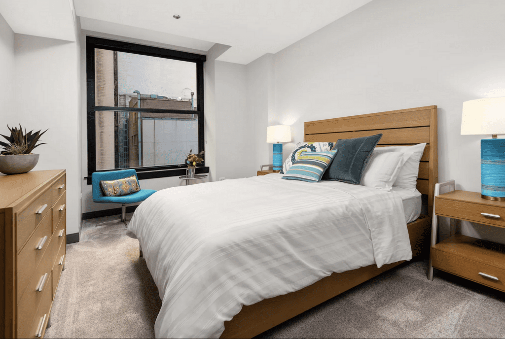 A modern bedroom with a wooden bed, striped cushions, two bedside tables with blue lamps, a dresser, and a teal chair. A window with a view of buildings is visible.