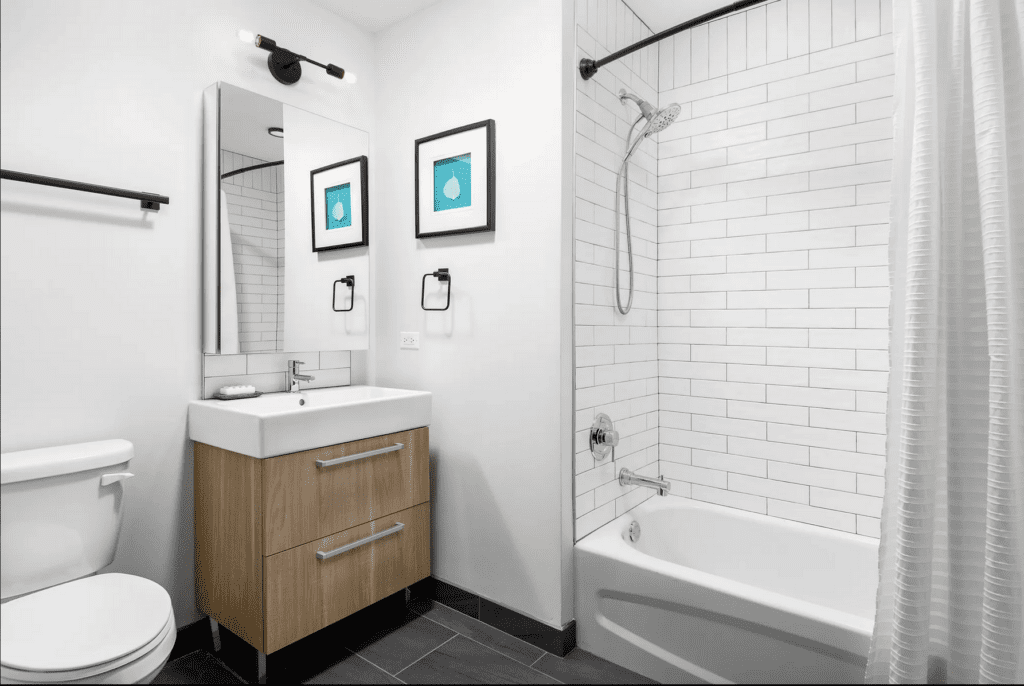 Modern bathroom with white tile walls, bathtub with shower, wood vanity with sink and mirror, toilet, two framed pictures, and black fixtures.