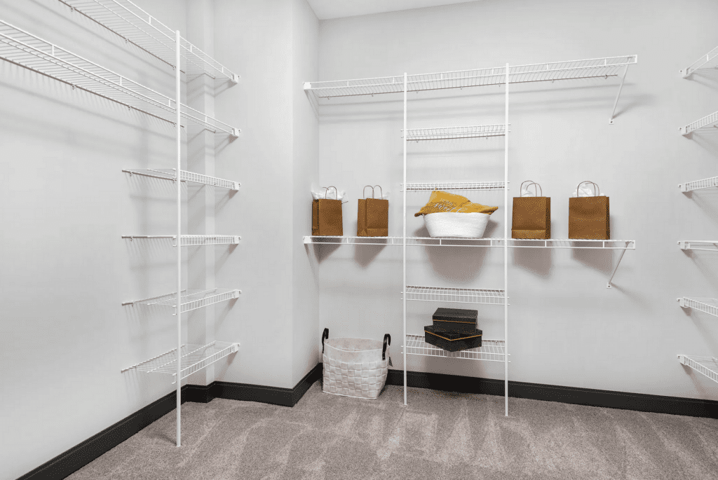 A walk-in closet with white wire shelves, containing several brown paper bags, a white basket with yellow fabric, a white woven basket, and a few black boxes. Carpeted floor and gray walls.
