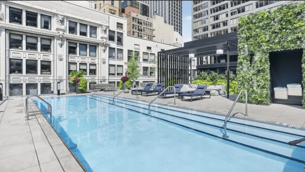 Rooftop pool area with lounge chairs, surrounded by tall buildings and a wall of greenery.