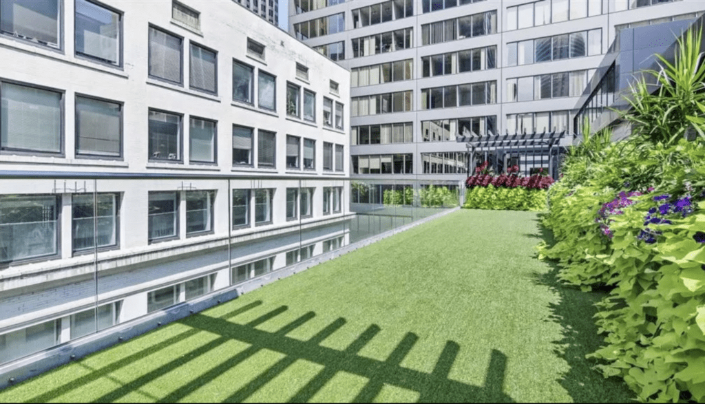 Rooftop garden with artificial grass, surrounded by modern buildings. Includes glass railings, potted plants, and a wooden pergola adorned with flowers.