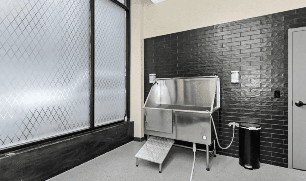 A pet washing station with a stainless steel tub, small stairs, black tiled walls, a frosted glass window, and wall-mounted soap holders.