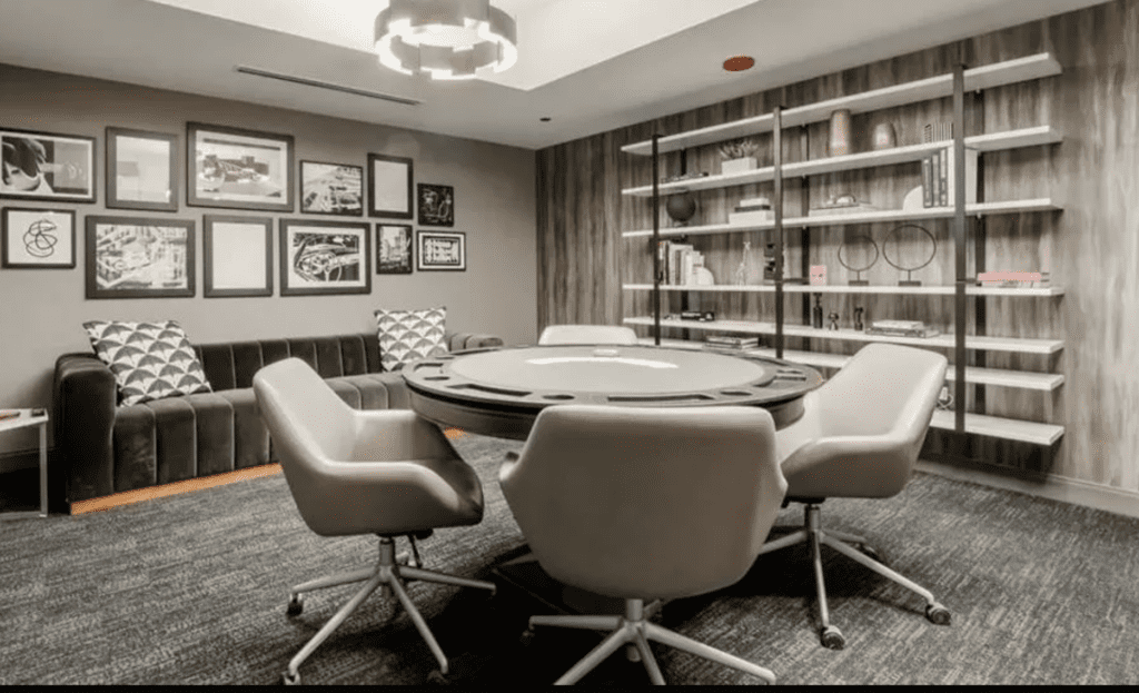 Modern room with a round poker table, four gray chairs, a gray sofa, wall art, and a bookshelf.